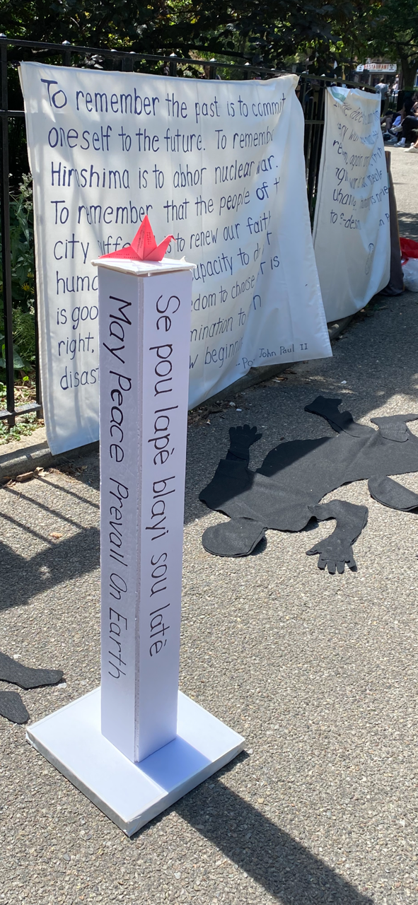 art installation depicting black shadows of women and children on the ground in front of peace post and banners describing stance on non-nuclear proliferation and anti-war message of the catholic church.
