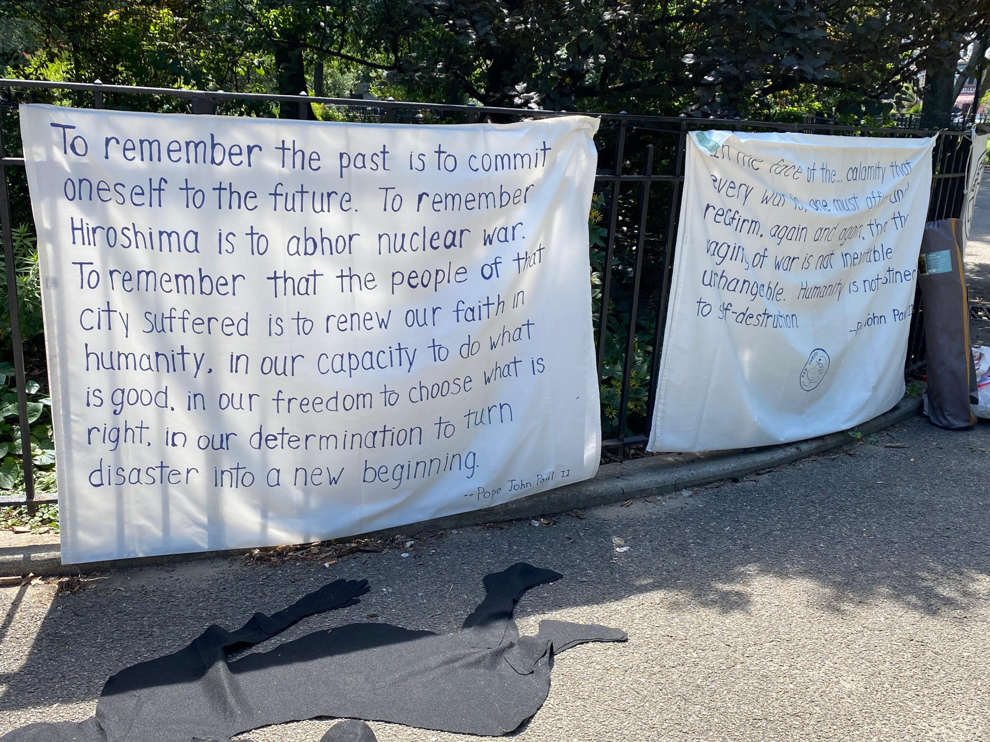 art installation depicting black shadows of women and children on the ground in front of peace post and banners describing stance on non-nuclear proliferation and anti-war message of the catholic church.