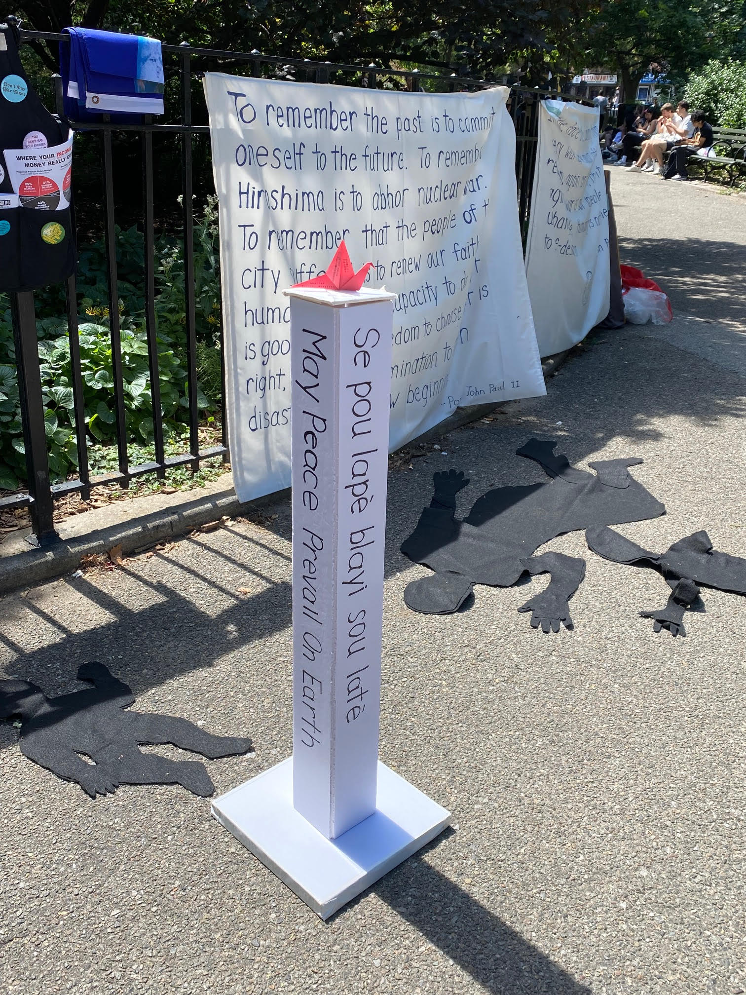 art installation depicting black shadows of women and children on the ground in front of peace post and banners describing Pax Christi stance on non-nuclear proliferation and anti-war message of the catholic church.